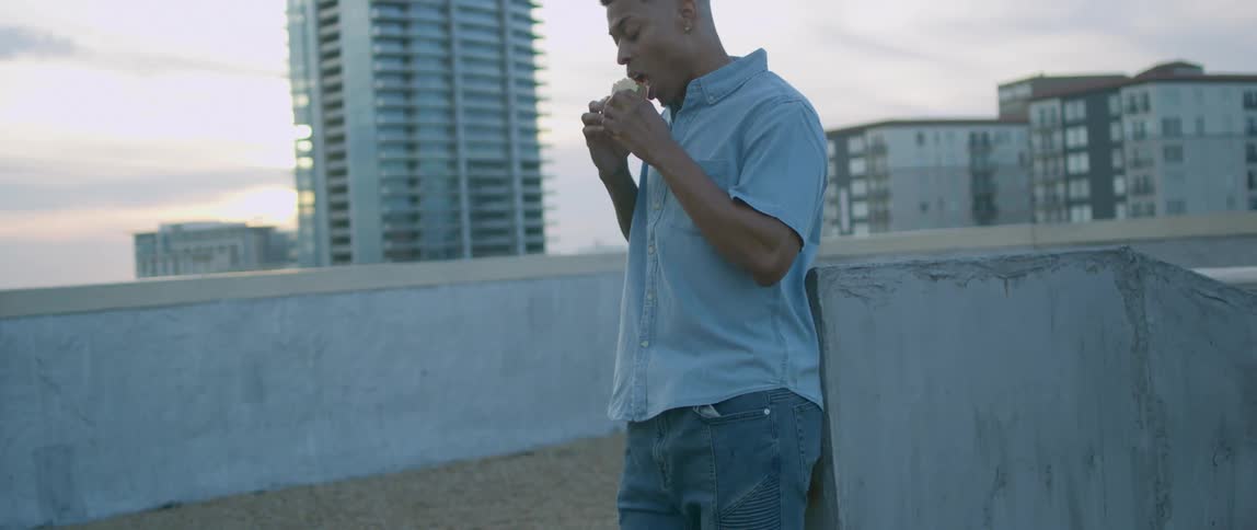 A black man eats a sandwich while standing on the roof of a building