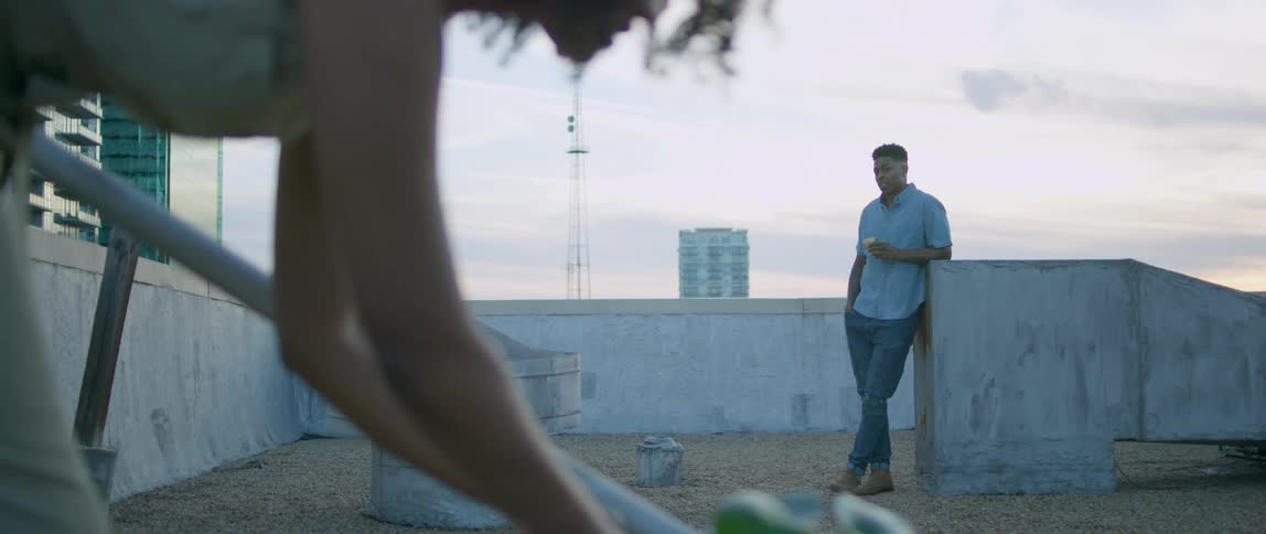 A man standing on a rooftop watches a woman water a plant