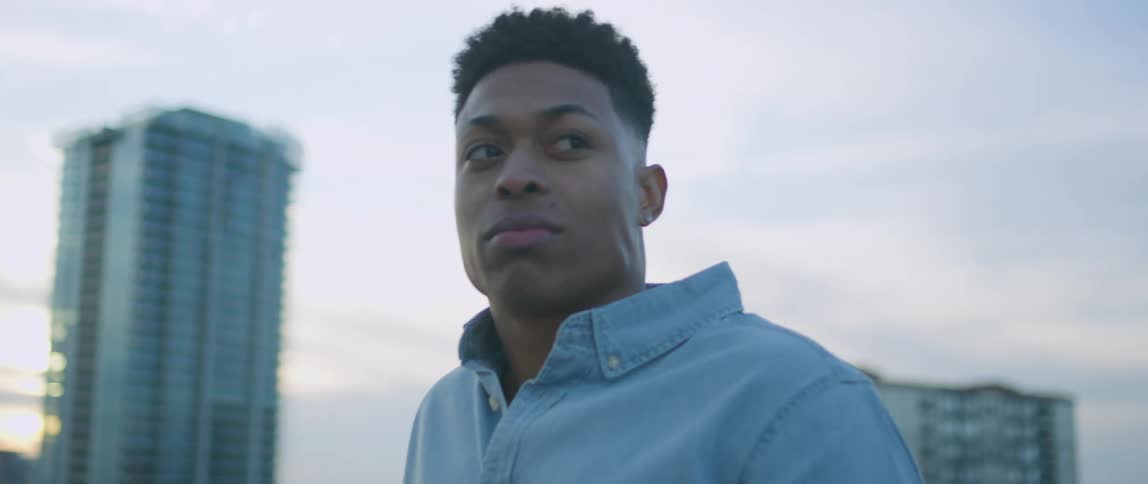 A young black man takes a bite of his sandwich before turning to smile at something and walking towards it out of frame