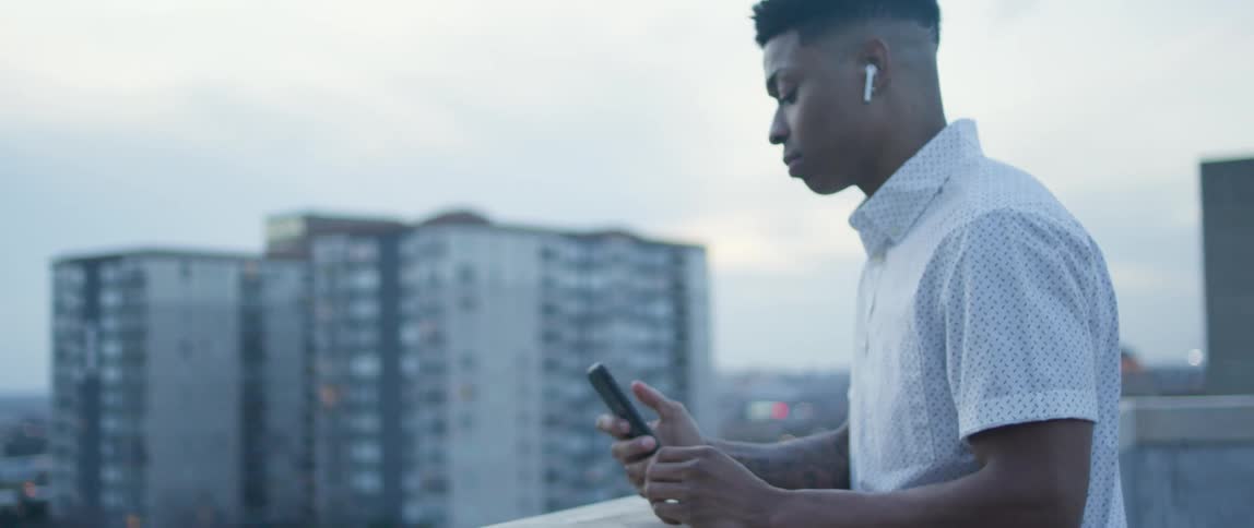 Male leans against the ledge of the balcony in a highrise building as he places an earbud into his right ear then cues the music on his cell phone and listens to music while nodding his head to the beat while looking out over the city