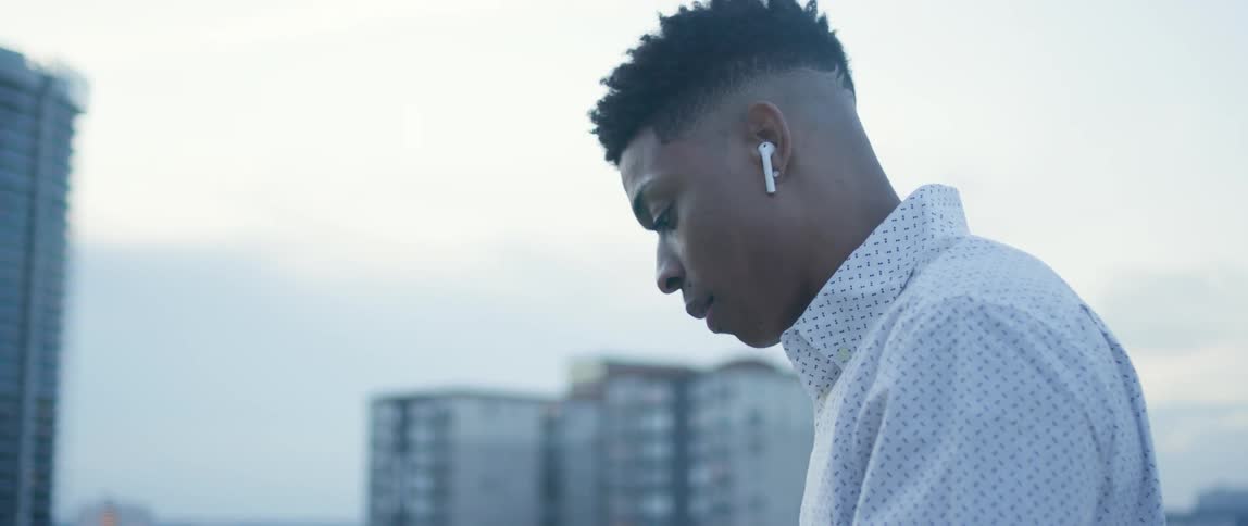 A black man puts an air pod into his ear while standing on a building rooftop