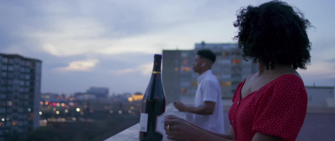 A woman on a roof top sets a bottle of wine and glasses down and then picks them up and goes over to a man on the roof top