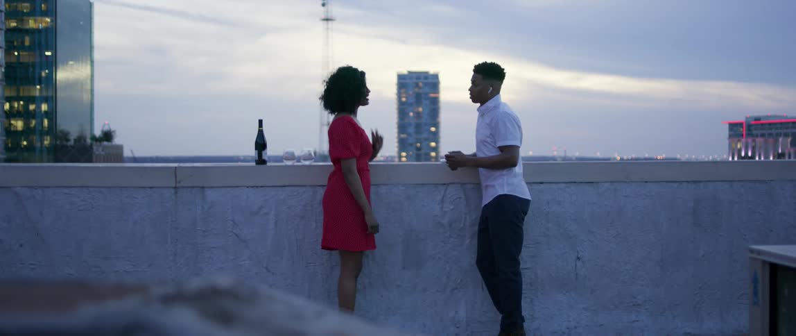A man and woman stand together on a city rooftop with a bottle of champagne