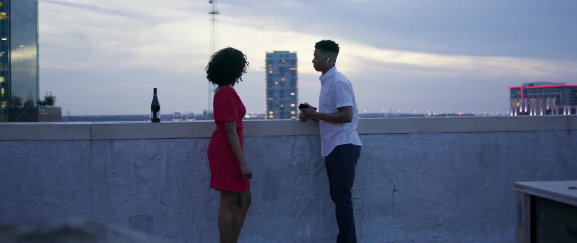 A black man and black woman stand on a city rooftop and talk and dance