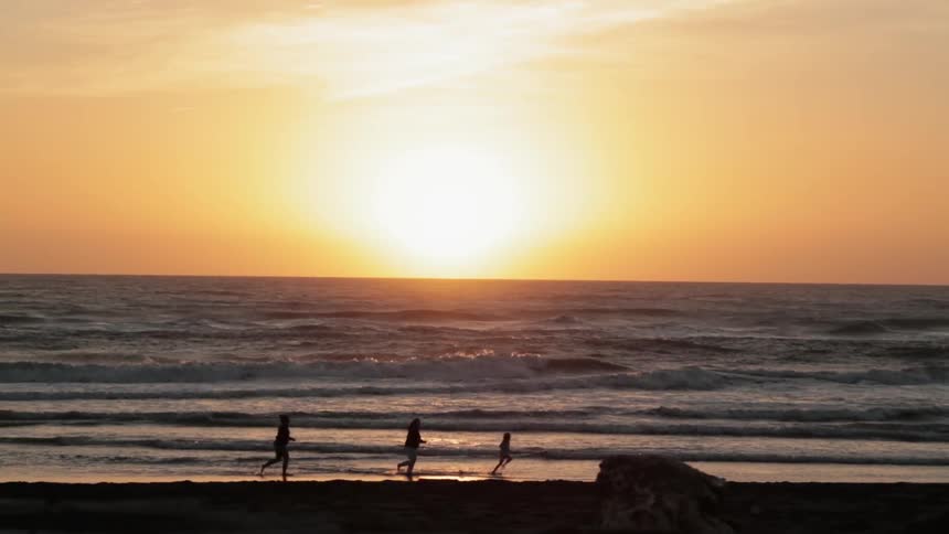 Three people run along the beach as the sun sets and waves roll in
