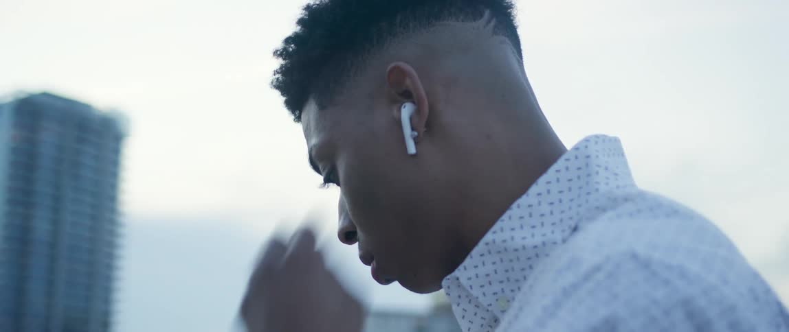 A black man stands on a rooftop messing with his smartphone and using an air pod