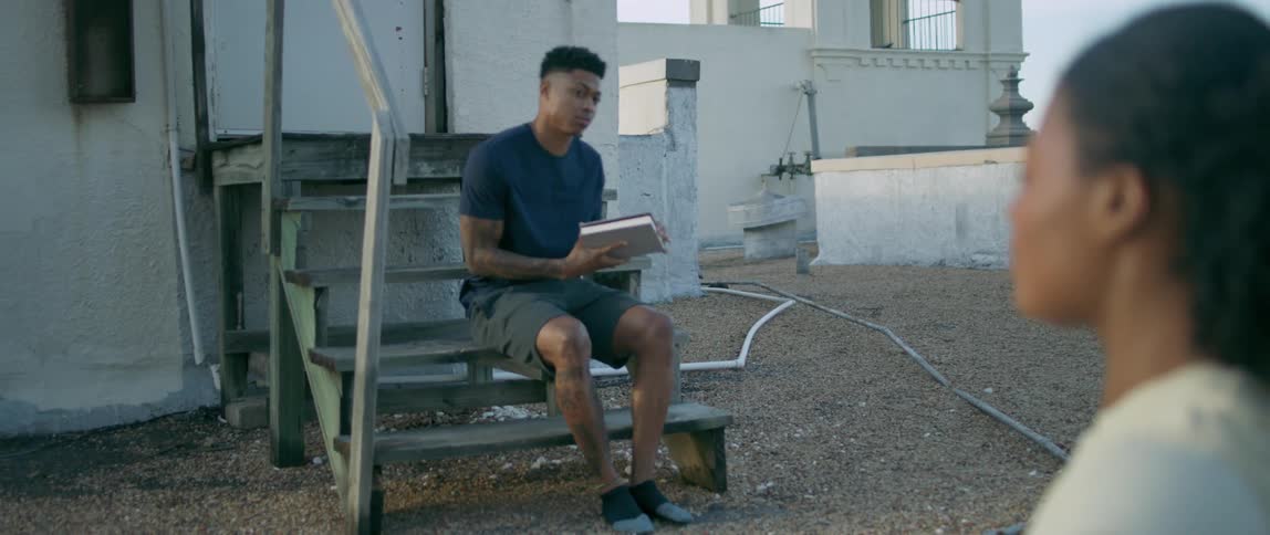 A young woman on a roof top meditates and a young black man comes and sits down on the stairs to read a book