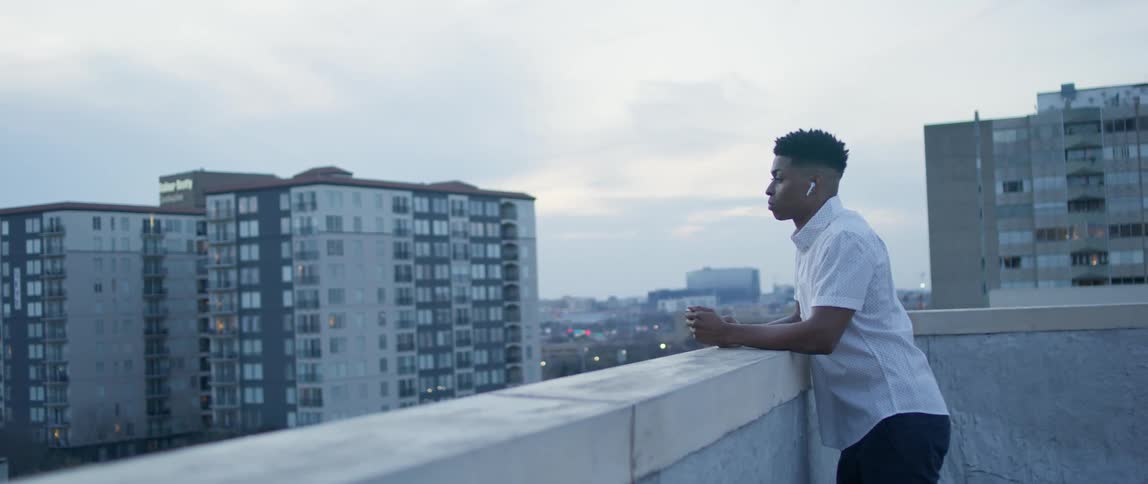 A young man stands on a city rooftop listening to earbuds