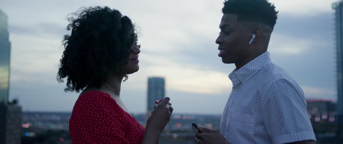 A man on a rooftop takes out an airpod and gives it to a woman and starts dancing
