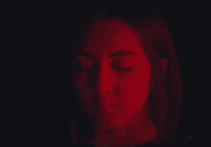 A white woman stands still with her eyes closed while bathed in red light