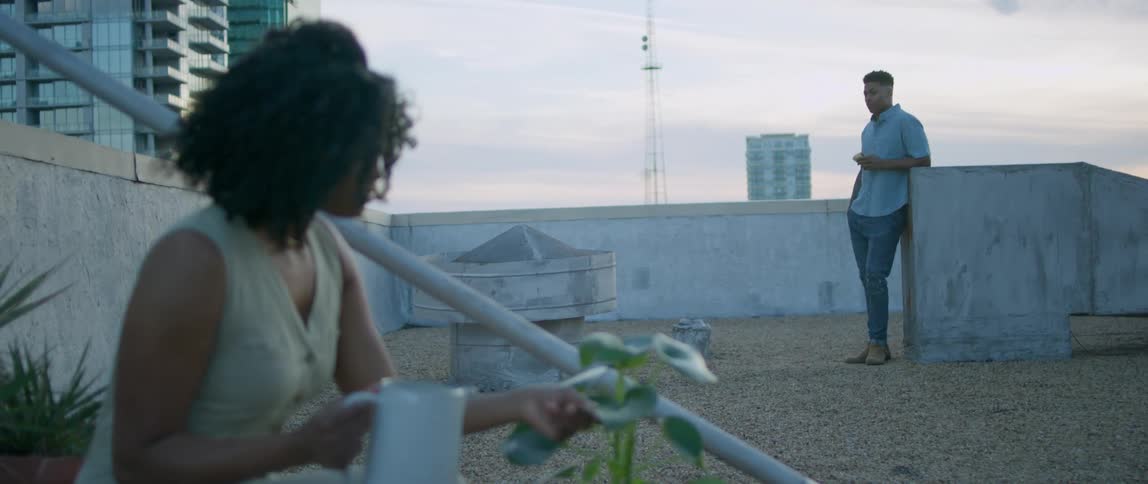 A black woman waters plants on a rooftop while a black man stands close by eating a sandwich