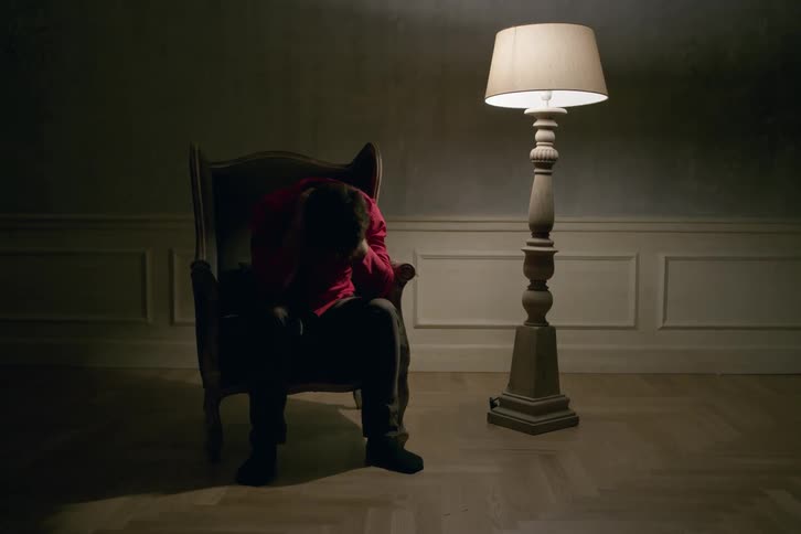 Depressed man sitting on a chair next to a lamp in a dimly lit room