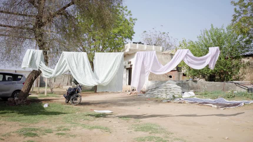Sheets are hung to dry on an outdoor clothing line outside a house over a dirt road