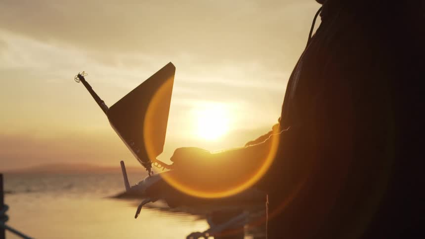 Man stands near the ocean silhouetted by sun as it nears the horizon and manipulates drone remote