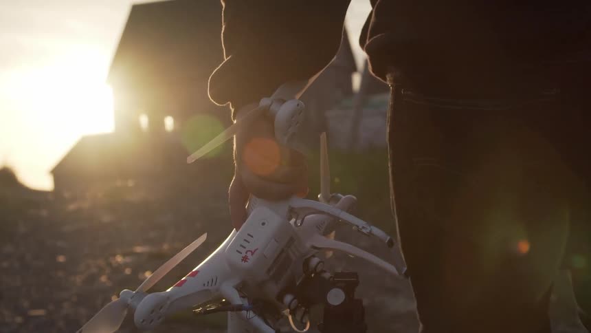 A white man carries a drone up a dirt path near a building