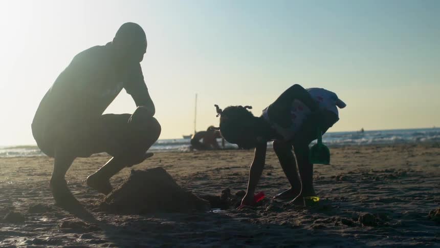 A father watching his daughter dig in the sand with plastic shovels on the beach