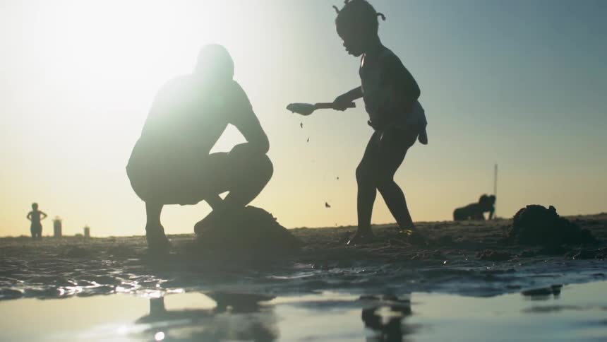 Father kneeling as daughter uses a shovel to scoop the wet sand to add to a sand pile she is making at the beach as the sun is setting