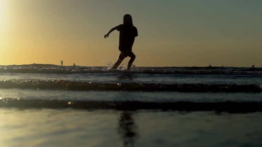 Woman silhouetted by sun runs across waves rolling onto beach as others ride their boards near horizon