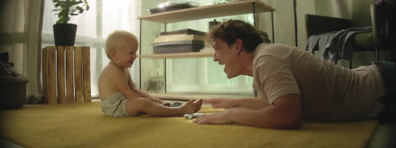 A white man lies on the floor with a baby and plays around with him