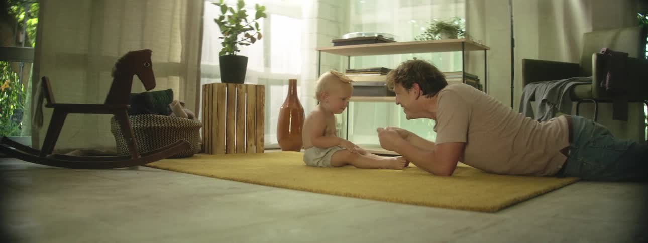 Father laying on his stomach on the floor in front of son while placing toys on babys head and laughing when the objects fall