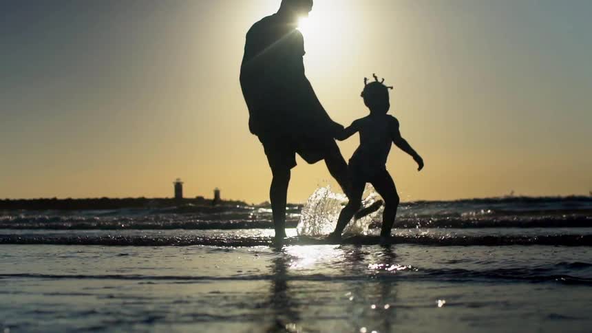 Father and daughter hold hands as they run and splash on the edge of the water of the ocean while at the beach at dusk