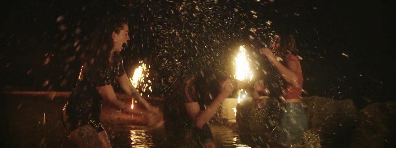Two women and a man splash each other and laugh in a shallow pond with sparkler torches on shore at night