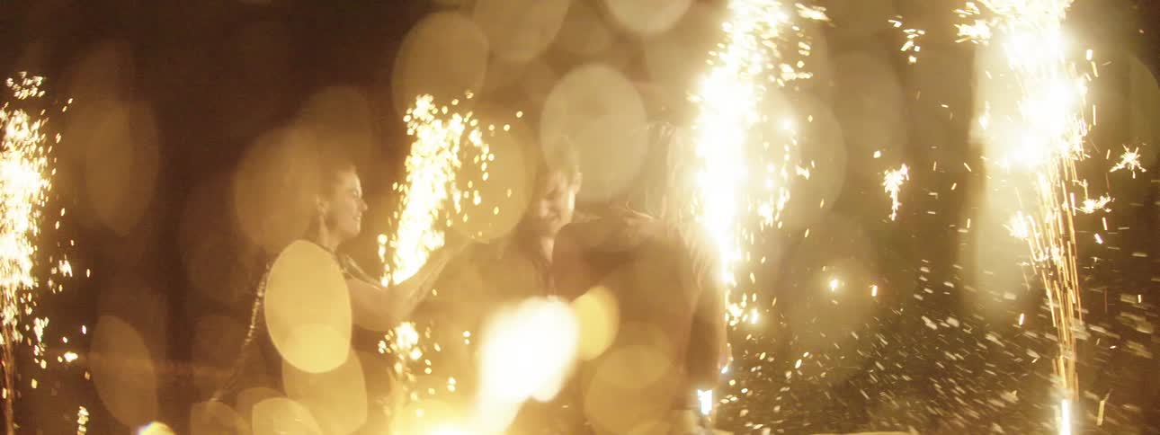 A girl jumps on a boys back as the three teens play in the water while flares burn