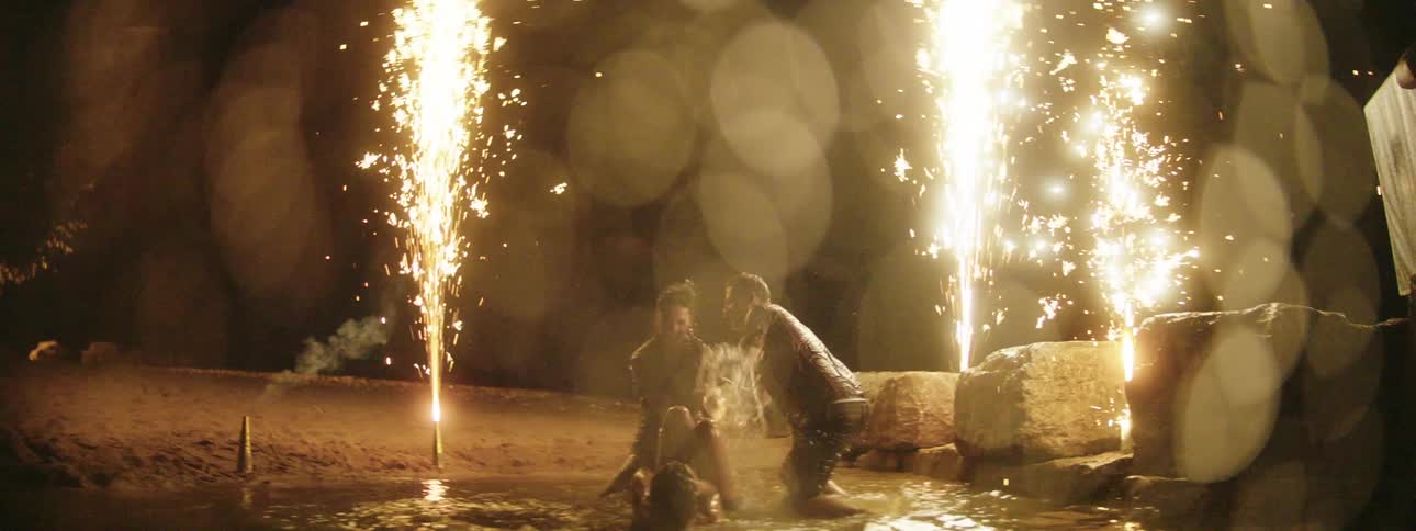 People play in water near beach as fireworks shoot sparks into the sky at night
