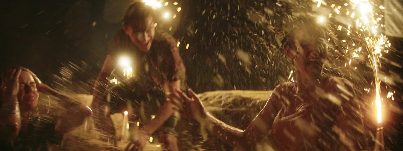 Three white teens frolic and splash in a lake while fireworks go off behind them