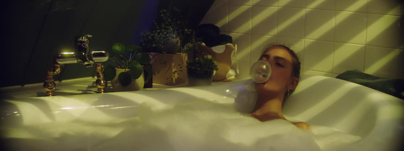 A young woman lies in a bath tub with her eyes closed and blows gum bubble