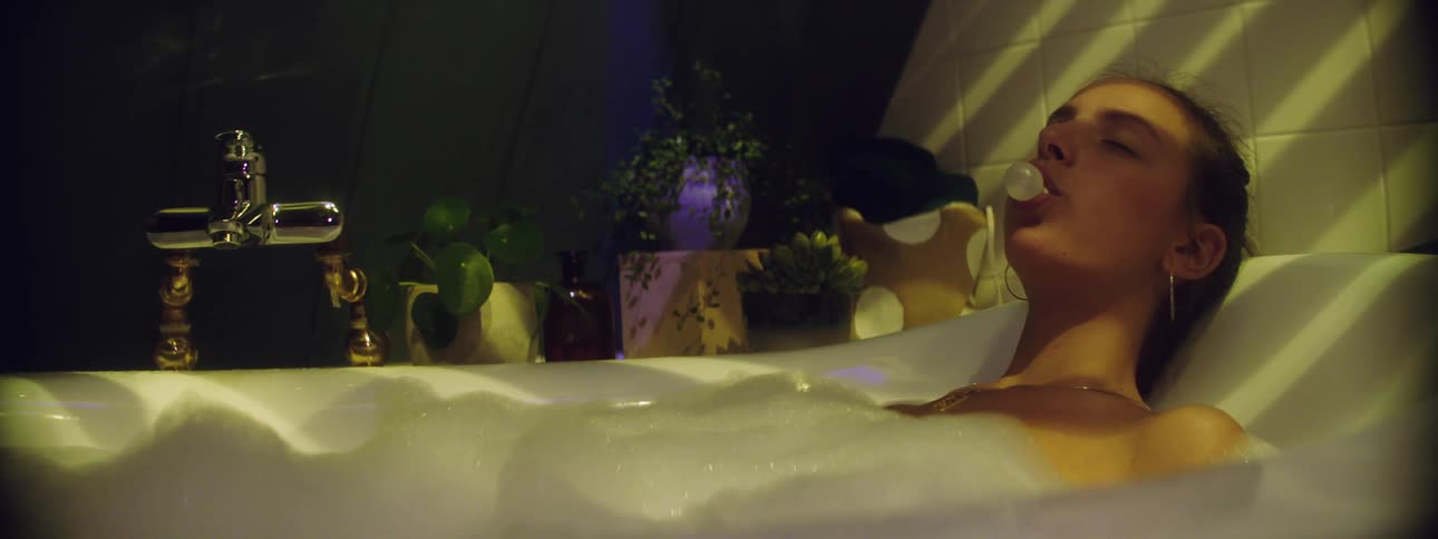 Female laying in a tub full of bubbles has her eyes closed as she chews on a piece of gum and blows a bubble