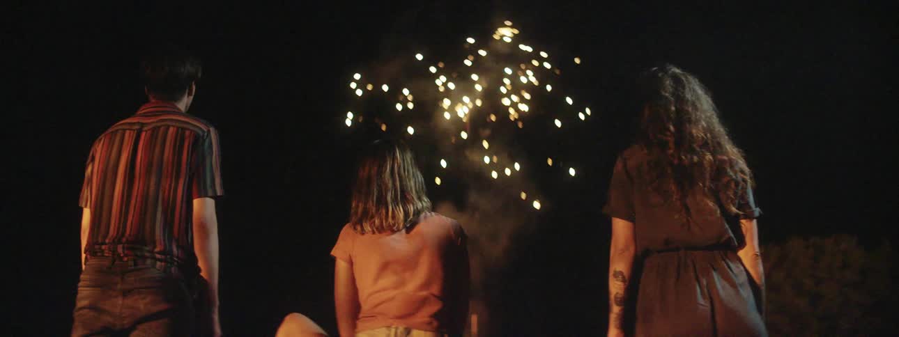 Three white people stand outdoors and watch fireworks pop in the sky