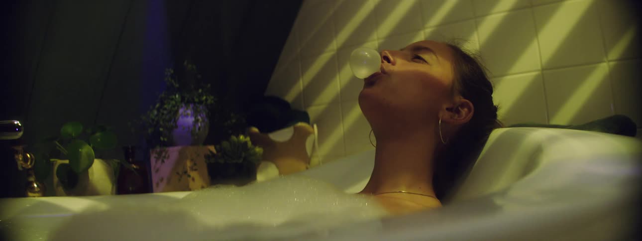 Woman chews gum and blows a bubble while taking a bubble bath in a bath tub