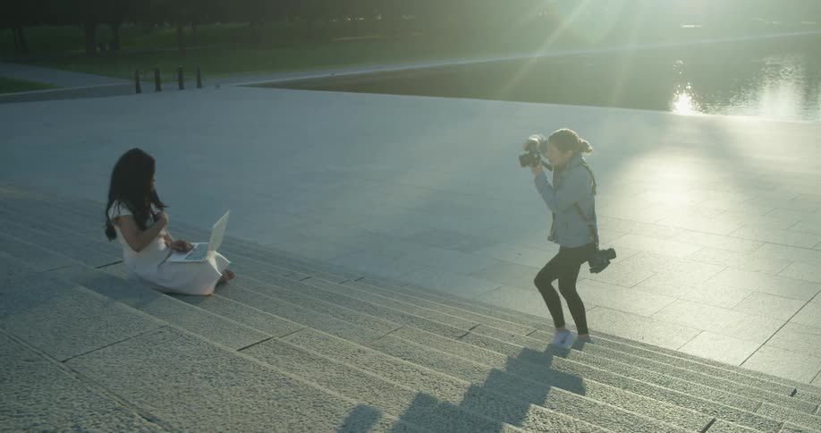 A white female photographer takes a picture of a woman using a laptop in a park