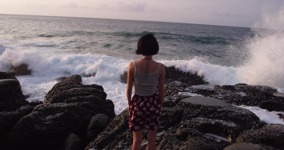 A young woman stands on a rocky coastline as the waves come in