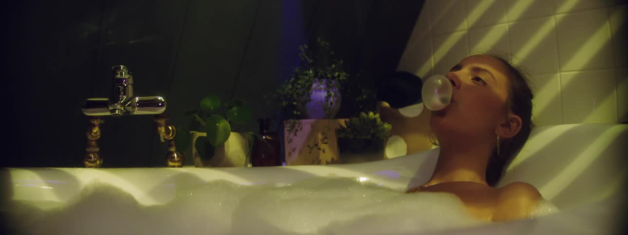 A young white woman sits in a bathtub blowing a bubble with gum