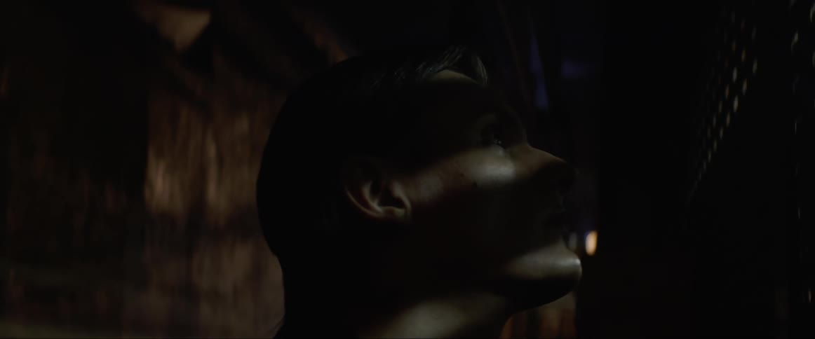 A man stands outside looking in through a grated window as the bright light casts shadow on his face