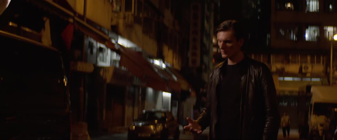 A man stands in a dark city street looking at his hand and his surroundings in confusion