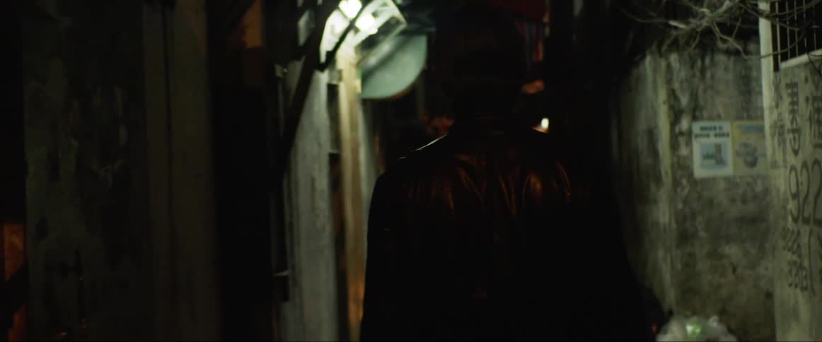 A white man in a jacket walks down a cramped alley in a city neighborhood