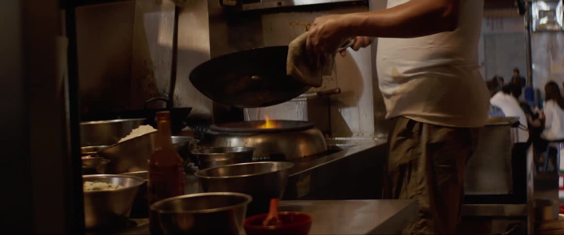 A person cooks in a restaurant kitchen as people sit in the restaurant at tables