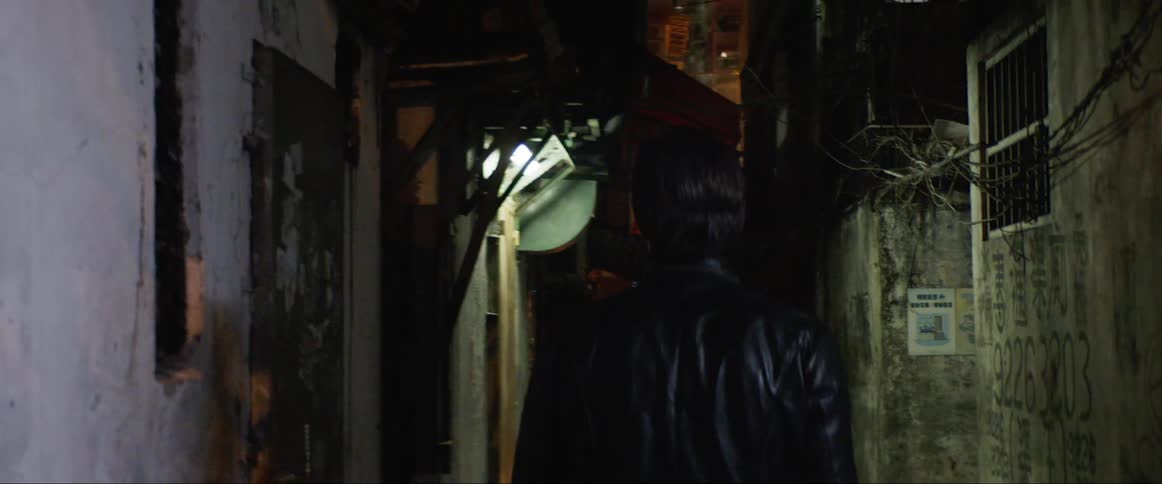 A white man walks down a dark alleyway and pauses to rub his hand on a wall