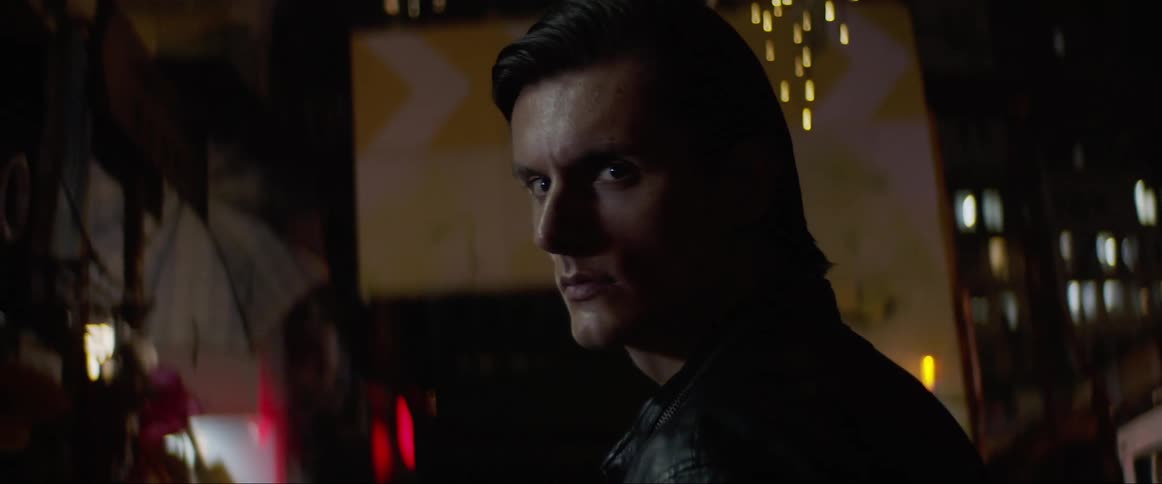 Man looks over his right shoulder as he stands in a room with a light flashing behind him