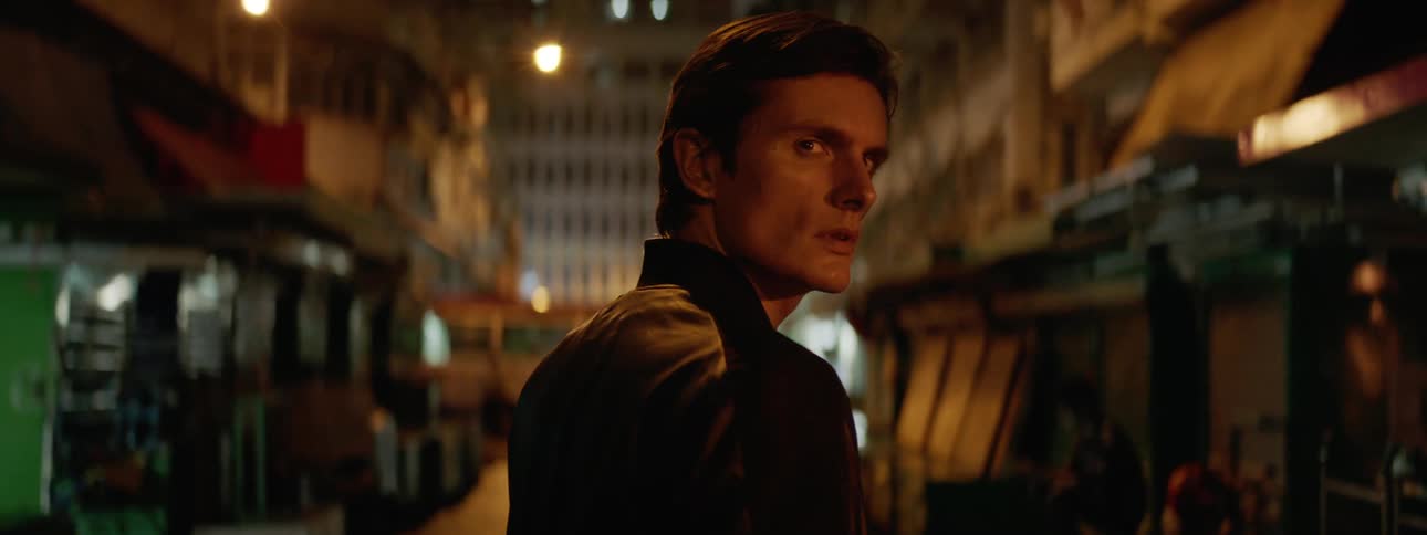 Man wearing leather jacket walks down narrow street between small shops turning to look behind