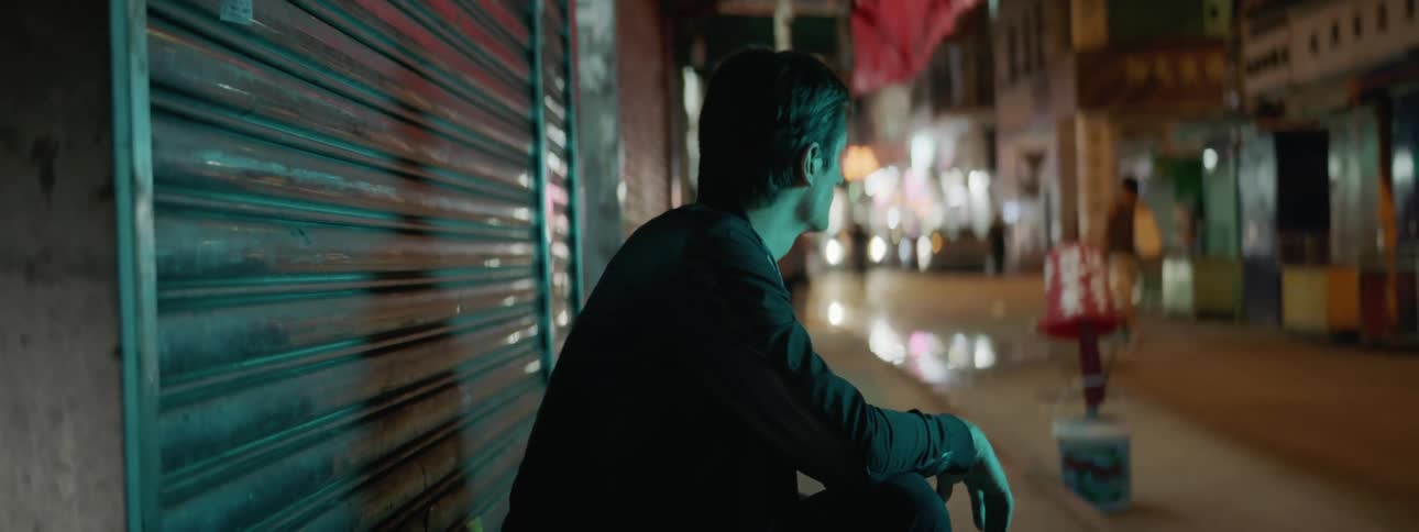 A man sits on a sidewalk with his arm resting on his knee watching people walk by on a city street at night