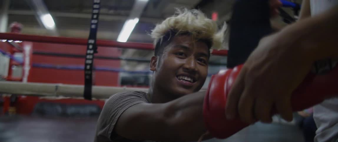 A boxer sits on the edge of a ring and smiles as a trainer wraps and puts on his glove