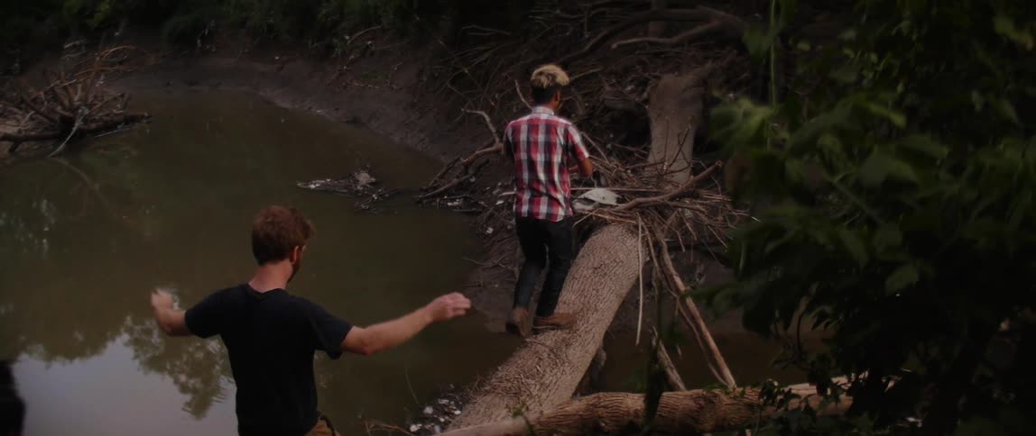 Three teenage boys walk across a tree trunk over a river while one carries a fishing pole