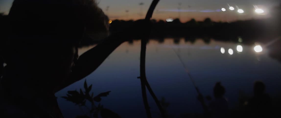 A young man speaking and looking over two children fishing in lake at night as he breaks twigs off a fallen tree branch