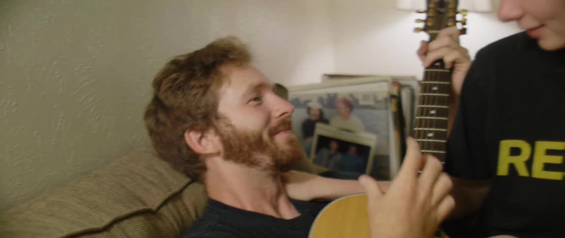 Female sits on males lap who is strumming on a guitar while sitting on the couch as they talk and laugh together