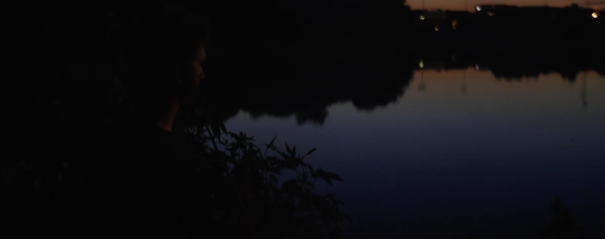 A white teenager stands on the shore of a lake at dusk