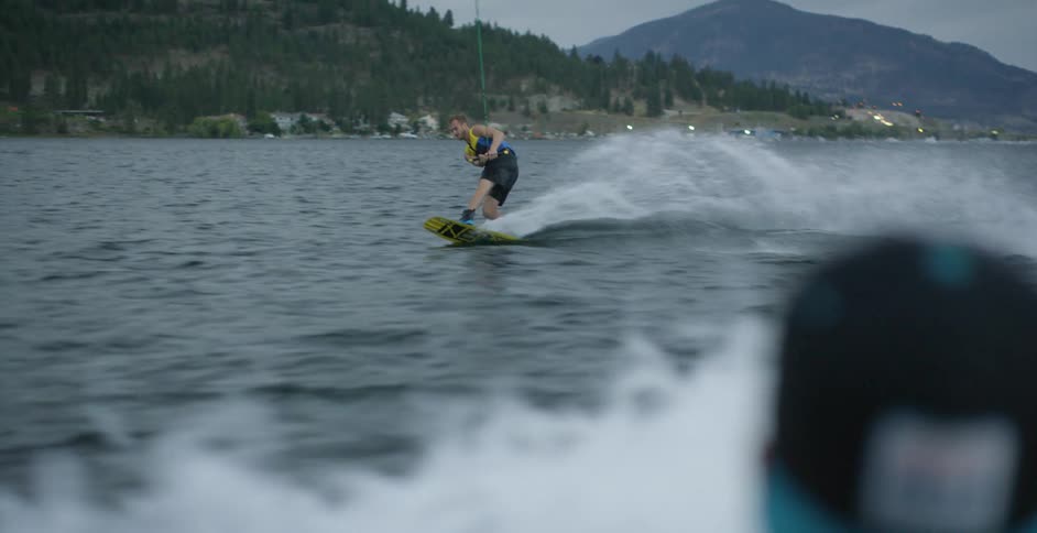 A man in life vest wakeboarding off the coast as another man in a baseball cap watches
